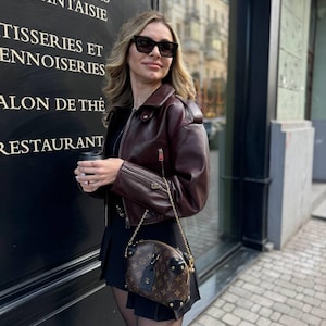 May include: A woman wearing a burgundy leather jacket, black sunglasses, and a black pleated skirt. She is carrying a brown handbag with a gold chain strap. The handbag has a brown monogram pattern and gold studs.