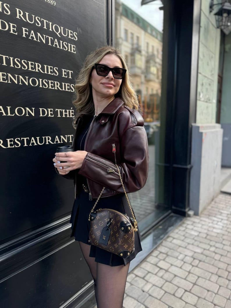 May include: A woman wearing a burgundy leather jacket, black sunglasses, and a black pleated skirt. She is carrying a brown handbag with a gold chain strap. The handbag has a brown monogram pattern and gold studs.