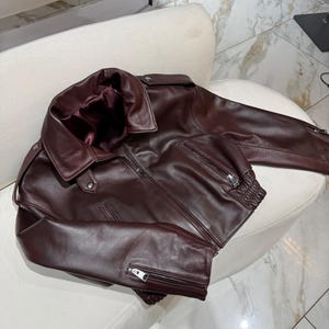 May include: A burgundy leather jacket with a wide collar and a zipper closure. The jacket has two front pockets with zippers and a ribbed hem. The jacket is displayed on a white chair.