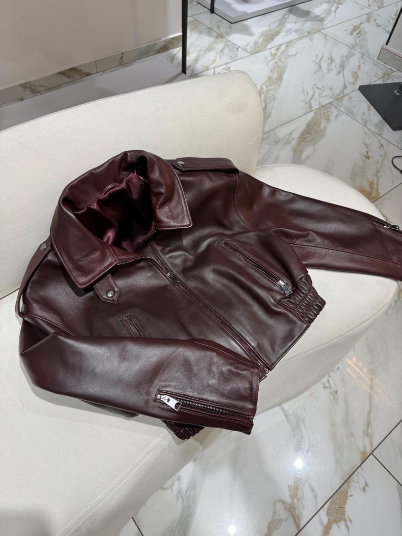 May include: A burgundy leather jacket with a wide collar and a zipper closure. The jacket has two front pockets with zippers and a ribbed hem. The jacket is displayed on a white chair.