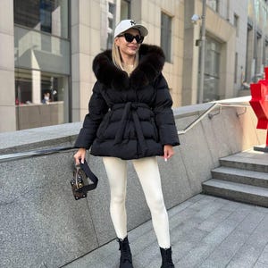 May include: A black puffer jacket with a fur-trimmed hood and a tie waist. The jacket is worn with white leggings and black combat boots. A white baseball cap and sunglasses complete the outfit. A small black handbag is held in the hand.