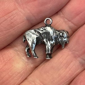 May include: A small, silver-toned buffalo charm with a loop for attaching to a necklace or bracelet. The charm is detailed with the buffalo's features, including its horns and fur. The charm is likely made of metal and is a miniature representation of the animal.