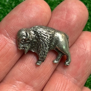 May include: A small, detailed silver-colored bison figurine. The miniature sculpture depicts the animal in a standing pose, with textured fur and horns. The bison is a symbol of the American West.