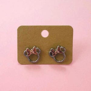 Puede incluir: Un par de pendientes de botón plateados con un diseño de lazo dorado. Los pendientes se muestran en una tarjeta marrón.