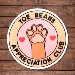 Peut inclure: Autocollant rond avec l'inscription "TOE BEANS APPRECIATION CLUB". Il présente une empreinte de patte de chat, deux cœurs rouges et un fond rose et beige. Le fond est une texture de bois.
