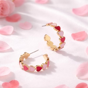 May include: Gold-coloured hoop earrings featuring alternating pink and red heart-shaped enamel designs. The earrings are displayed on a white surface scattered with pink rose petals, creating a romantic and feminine aesthetic.