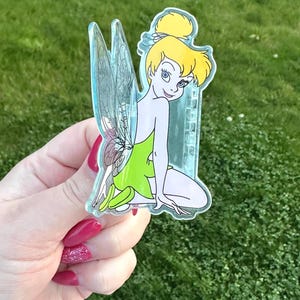 May include: A clear acrylic charm featuring a cartoon fairy with blonde hair in a bun, wearing a green dress and transparent wings. The fairy is kneeling and smiling. The charm is held against a background of green grass.
