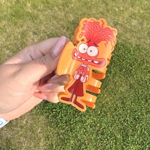May include: An orange cartoon character-shaped hair clip with red hair, a red mouth, and a white shirt with a red skirt. The clip is held against a background of green grass.