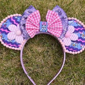 May include: A headband featuring two circular ears with a blue patterned fabric, purple wavy trim, and pink pom-pom edges. A central bow combines purple sequins with a pink and white gingham fabric, featuring a small square charm of a cartoon duck.
