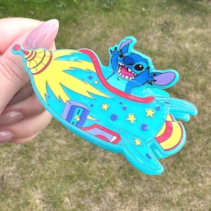 Puede incluir: Un personaje de dibujos animados azul en un cohete. El cohete es azul claro con detalles amarillos y rojos, y el personaje está sonriendo. La imagen es un diseño colorido y caprichoso.