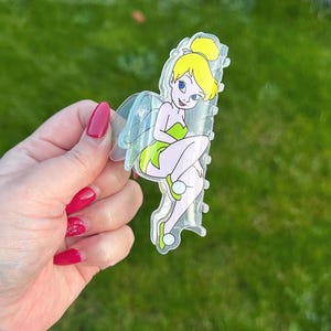 May include: A clear acrylic hair clip featuring a cartoon image of Tinkerbell. The clip has a yellow bun, green dress, and translucent wings. The background is a blurred green lawn.