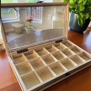 May include: Wooden jewelry box with a clear glass top and multiple compartments. The box is open, revealing the interior with small, square storage spaces. The wood has a natural grain and a light brown finish. The box is on a wooden surface.