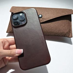 Peut inclure: Un étui de téléphone en cuir marron foncé et une pochette en feutre marron assortie. L'étui a une texture lisse et un module caméra à trois objectifs. La pochette a une fermeture à rabat avec un bouton-pression argenté.