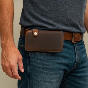 May include: A brown leather phone case with a snap closure is attached to a brown leather belt. The belt is worn with blue jeans. The case is rectangular and appears to be designed to hold a smartphone.
