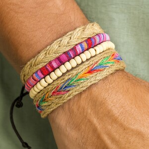 May include: A stack of woven bracelets in natural tones and colorful accents. The bracelets include a braided jute band, a striped fabric band with pink and red hues, a strand of wooden beads, and a band with embroidered rainbow details. A black cord is visible.