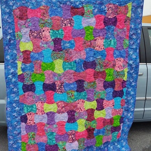 May include: A colorful handmade quilt with a repeating pattern of connected shapes in shades of pink, purple, blue, and green. The quilt has a blue border with a subtle floral design, and is likely made of cotton fabric.