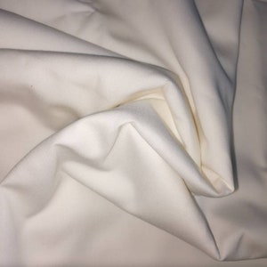 Könnte beinhalten: Cremefarbener Stoff mit sanften Falten und Schatten. Das Material hat ein glattes, undurchsichtiges Aussehen mit leichtem Glanz. Das Bild zeigt die Textur und den Fall des Stoffes, ideal für Kleidung oder Heimtextilien.