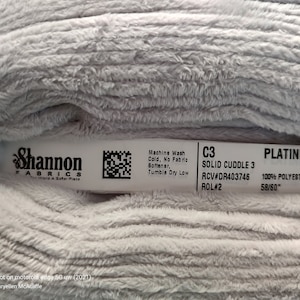 May include: A white fabric swatch labeled "Platinum" with the text "Solid Cuddle 3" and "100% Polyester 58/60" printed on it. The fabric is a soft, plush material.