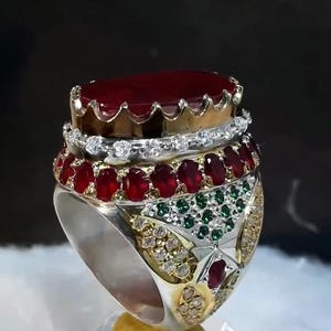 May include: A large, ornate ring featuring a large, oval-cut, deep red gemstone at the top. The ring has multiple layers of gemstones, including smaller red and green stones, and is set in a silver and gold metal band. The ring is highly detailed and appears to be handcrafted.