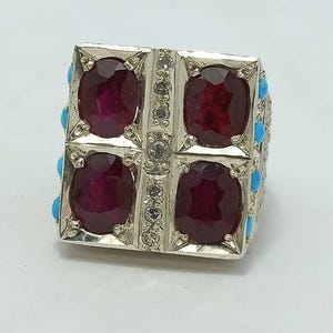 May include: A silver ring with four red ruby stones and a white diamond in the center. The ring is surrounded by small turquoise stones.