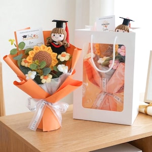 Puede incluir: Un ramo de ganchillo con un girasol, pequeñas flores blancas y una muñeca con birrete negro y borla roja. El ramo está envuelto en papel naranja con una cinta blanca impresa con el texto JUST FOR YOU.