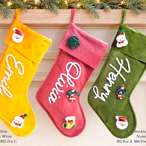 Könnte beinhalten: Drei Weihnachtssocken aus Samt in Gelb, Pink und Salbeigrün, jeweils mit einem weiß gestickten Namen: Emily, Olivia und Henry. Die Socken sind mit kleinen Filz-Weihnachtsmotiven wie einem Weihnachtsmann, einem Baum und einem Schneemann verziert.