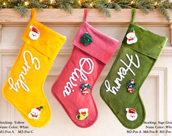 May include: Three velvet Christmas stockings in yellow, pink, and sage green, each featuring a white embroidered name: Emily, Olivia, and Henry. The stockings are decorated with small felt holiday icons including Santa, a tree, and a snowman.