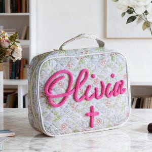 Puede incluir: Un bolso acolchado con estampado floral y asa, con el nombre "Olivia" en hilo rosa brillante. Una cruz rosa está debajo del nombre. El bolso está sobre una superficie de mármol.