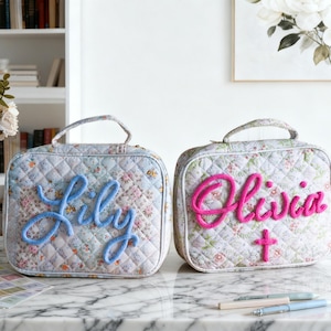 Könnte beinhalten: Zwei gesteppte Kosmetiktaschen mit Blumenmustern. Eine Tasche hat "Lily" in Blau gestickt, die andere "Olivia" in Pink mit einem rosa Kreuz. Beide haben Tragegriffe und stehen auf einer Marmoroberfläche.