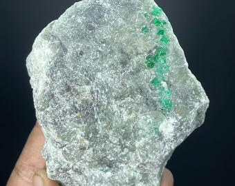 May include: A rough-cut, gray-green stone with vibrant green emerald crystals embedded throughout. The natural rock formation has a textured surface and irregular shape, showcasing the raw beauty of the mineral specimen. The background is a solid black.