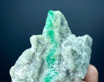 May include: A rough, irregularly shaped, light gray rock with a vibrant green vein running through it. The rock has a textured surface and is held in a person's hand. The green color is concentrated in a central band.
