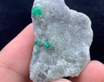 May include: A rough, light green mineral specimen with several small, vibrant green emerald crystals embedded within. The stone has a textured surface and a natural, irregular shape, showcasing the raw beauty of the gemstone.