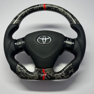 Könnte beinhalten: Ein Toyota-Lenkrad mit schwarzem Ledergriff, perforierten Seiten und einer Oberfläche aus geschmiedetem Carbon an den oberen und unteren Abschnitten. Ein vertikaler roter Streifen ist oben zentriert und ein GR-Logo ist unten zentriert.