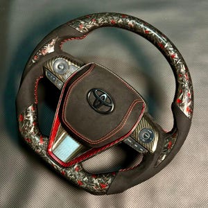 以下が含まれることがあります： 黒いスエードグリップと赤いステッチが施されたカスタムカーのステアリングホイール。ホイールは、外側のリムにカーボンファイバーと赤のパターンデザインが施されています。黒いトヨタのエンブレムがステアリングホイールの中央に配置されています。