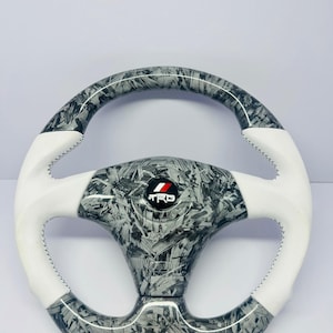 May include: A steering wheel with a black and gray pattern and white accents. The wheel has a TRD logo in the center and white stitching. The wheel is round and appears to be made of a composite material.