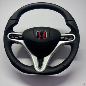 Könnte beinhalten: Schwarzes Lederlenkrad mit roten Nähten und silberner Mitte. Die Mitte hat eine schwarze Blende mit dem roten Honda-Logo. Das Lenkrad verfügt über integrierte Bedienelemente für Tempomat und andere Funktionen.