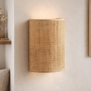 Pode incluir: Arandela de parede cilíndrica em rattan tecido, montada numa parede clara. A arandela emite um brilho quente, criando um ambiente suave. O design apresenta um padrão natural e texturizado, adicionando um toque de elegância rústica ao espaço.