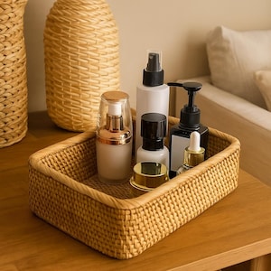 Könnte beinhalten: Ein rechteckiges Tablett aus geflochtenem Rattan, gefüllt mit verschiedenen Kosmetikflaschen und -behältern. Die Flaschen sind weiß, schwarz und goldfarben, mit Sprüh- und Pumpvorrichtungen. Das Tablett steht auf einer Holzoberfläche.