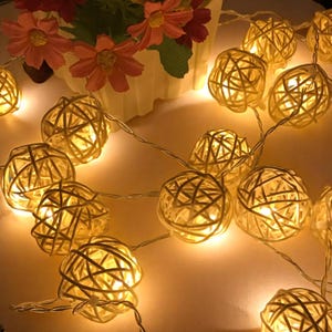 Può includere: Stringa di luci con sfere in rattan bianco caldo. Ogni sfera è illuminata, creando un bagliore soffuso. Le luci sono disposte su una superficie chiara, con un vaso di fiori rosa sullo sfondo.