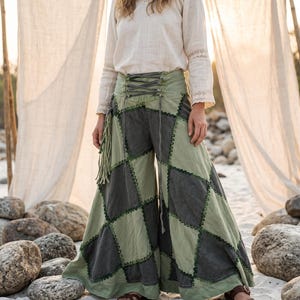 May include: A long-sleeved cream-colored blouse with lace trim and wide-leg pants featuring a patchwork diamond pattern in sage green and charcoal gray. The pants have a high-waisted lace-up front closure, decorative fringe, and dark green zigzag stitching along the seams.