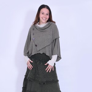 May include: An olive green layered skirt and matching poncho-style top. The skirt has a tiered design with textured fabric and a fringed hem. The top features button details and a draped neckline. The outfit is paired with neutral-toned sandals.