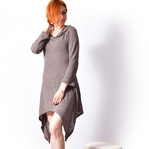 May include: A woman in a long-sleeved, hooded dress with an asymmetrical hem in a muted brown shade. She wears black boots. Beside her is a small, round stool with a cream top and a black and white patterned skirt.