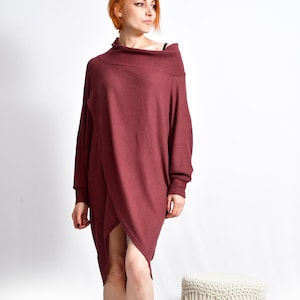 May include: A burgundy-coloured, long-sleeved dress with an asymmetrical hemline. The dress has a draped neckline and a wrap-style design. The model is wearing brown sandals. A small, round, macrame stool with wooden legs is to the right.