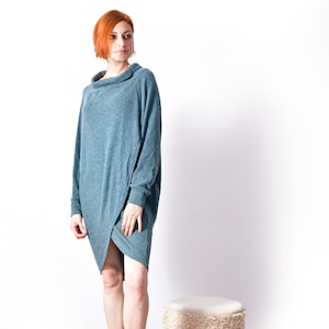 May include: A teal-coloured, long-sleeved dress with a cowl neck and asymmetrical hemline. The dress is worn with black ankle boots. A small, round stool with a cream-coloured top and a black and white patterned band sits on a woven rug.