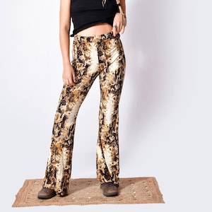 Puede incluir: Un top negro sin mangas y pantalones acampanados con un estampado de piel de serpiente en tonos marrón, beige y crema. Los pantalones tienen cintura alta y pernera ancha. El conjunto se lleva con zapatos marrones. La modelo está de pie sobre una pequeña alfombra tejida.