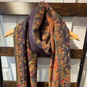 May include: A patterned scarf in shades of purple, orange, yellow, and red hangs on a wooden hanger. The scarf features a paisley design and has a fringe at the ends. The scarf is displayed on a wooden ladder.
