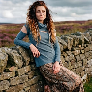 May include: A woman wearing a teal long-sleeved top with cut-out shoulders and a patterned skirt, leaning against a stone wall. Her long, wavy hair is a mix of brown and auburn. The background features a field of purple flowers under a cloudy sky.