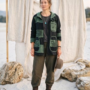 May include: A black zip-up hooded jacket featuring olive green panels with horizontal pleated detailing, worn over a dark gray t-shirt and olive green trousers, paired with brown leather boots and a brown felt hat held in one hand.