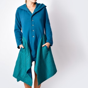 Pode incluir: Um casaco com capuz azul-petróleo com mangas compridas e fecho de botões. O casaco apresenta grandes bolsos azul-petróleo com um padrão texturizado. A modelo usa botas castanhas. É uma peça de vestuário única.