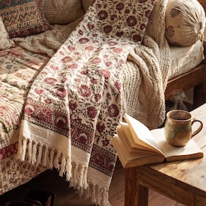 Puede incluir: Un espacio de vida acogedor con una manta estampada floral, cojines y una manta de punto. Una mesa auxiliar de madera sostiene un libro abierto y una taza de cerámica. Botas de cuero marrón descansan en el suelo de madera.